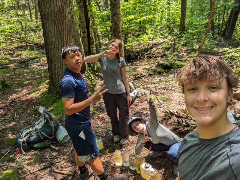 The image shows four people in a forest setting. One person is taking a selfie, smiling at the camera. To the left, a person is making a peace sign. Behind them, another person is also making a peace sign. In the foreground, there are several containers on the ground. A backpack is visible on the left side of the image.